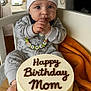 baby, infant, cake, birthday_cake, hat, onesie, crib, blanket, orange_blanket, wooden_table, indoor, window, hand, face, eyes, clothing, celebration, food, dessert, person