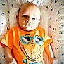 baby, infant, child, orange_tshirt, dinosaur_graphic, sunglasses, mickey_mouse_pattern, blanket, lying_down, cute, portrait, young_child, serious_expression, indoor, soft_lighting, hand, fist, head, face, toddler