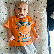 Enoan a rejoint le concours — aidez-le/la à gagner de superbes lots ! baby, infant, smiling, orange_tshirt, cartoon_dinosaur, sunglasses_graphic, shorts, blue_shoes, lying_down, mickey_mouse_pattern, cushion, child, happy, cute, indoors, playful, bright_colors, soft_surface, portrait, toddler