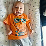baby, infant, smiling, orange_tshirt, cartoon_dinosaur, sunglasses_graphic, shorts, blue_shoes, lying_down, mickey_mouse_pattern, cushion, child, happy, cute, indoors, playful, bright_colors, soft_surface, portrait, toddler