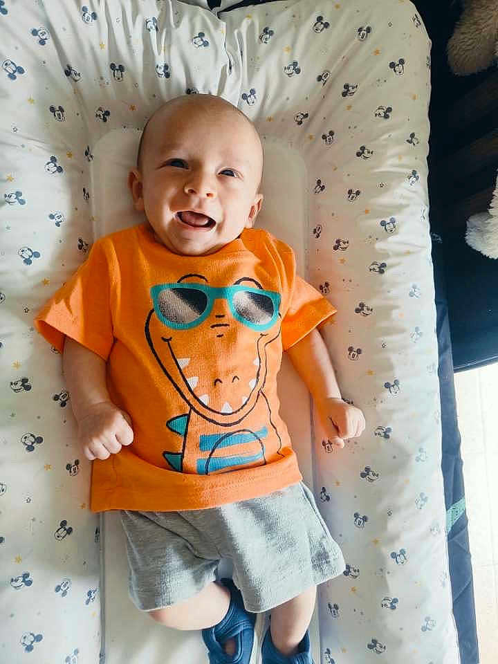 Enoan a rejoint le concours — aidez-le/la à gagner de superbes lots ! baby, infant, smiling, orange_tshirt, cartoon_dinosaur, sunglasses_graphic, shorts, blue_shoes, lying_down, mickey_mouse_pattern, cushion, child, happy, cute, indoors, playful, bright_colors, soft_surface, portrait, toddler