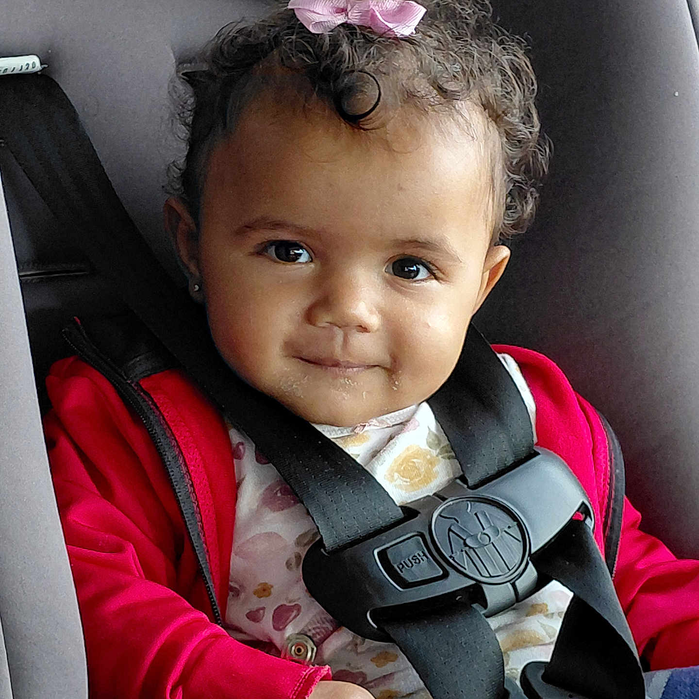 Selena joined the competition — help win amazing prizes! baby, car_interior, car_seat, child, child_safety, curly_hair, cute, face, floral_shirt, indoor, person, pink_bow, portrait, red_jacket, safety_harness, seat, seatbelt, smile, toddler, young_child