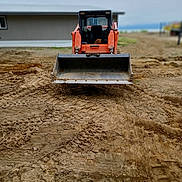 Junior is registered to the contest to win money with this photo: dog, bulldozer, construction_site, dirt, earth, vehicle, orange, window, building, sky, tire_tracks, outdoor, machine, pet, small_dog, seat, ground, calm, curious, worksite