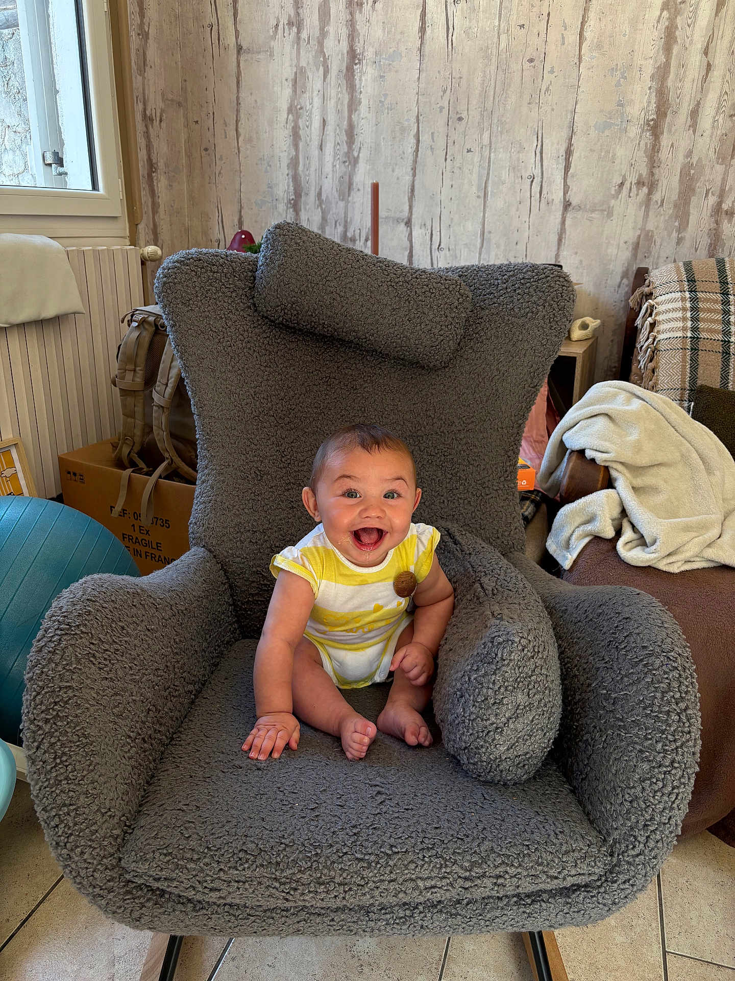 Pablo participe au concours pour gagner de l'argent avec cette photo : baby, child, smiling, happy, armchair, gray, plush, indoor, cozy, wooden_wall, window, floor, home, cushion, sitting, person, infant, clothing, furniture, radiator