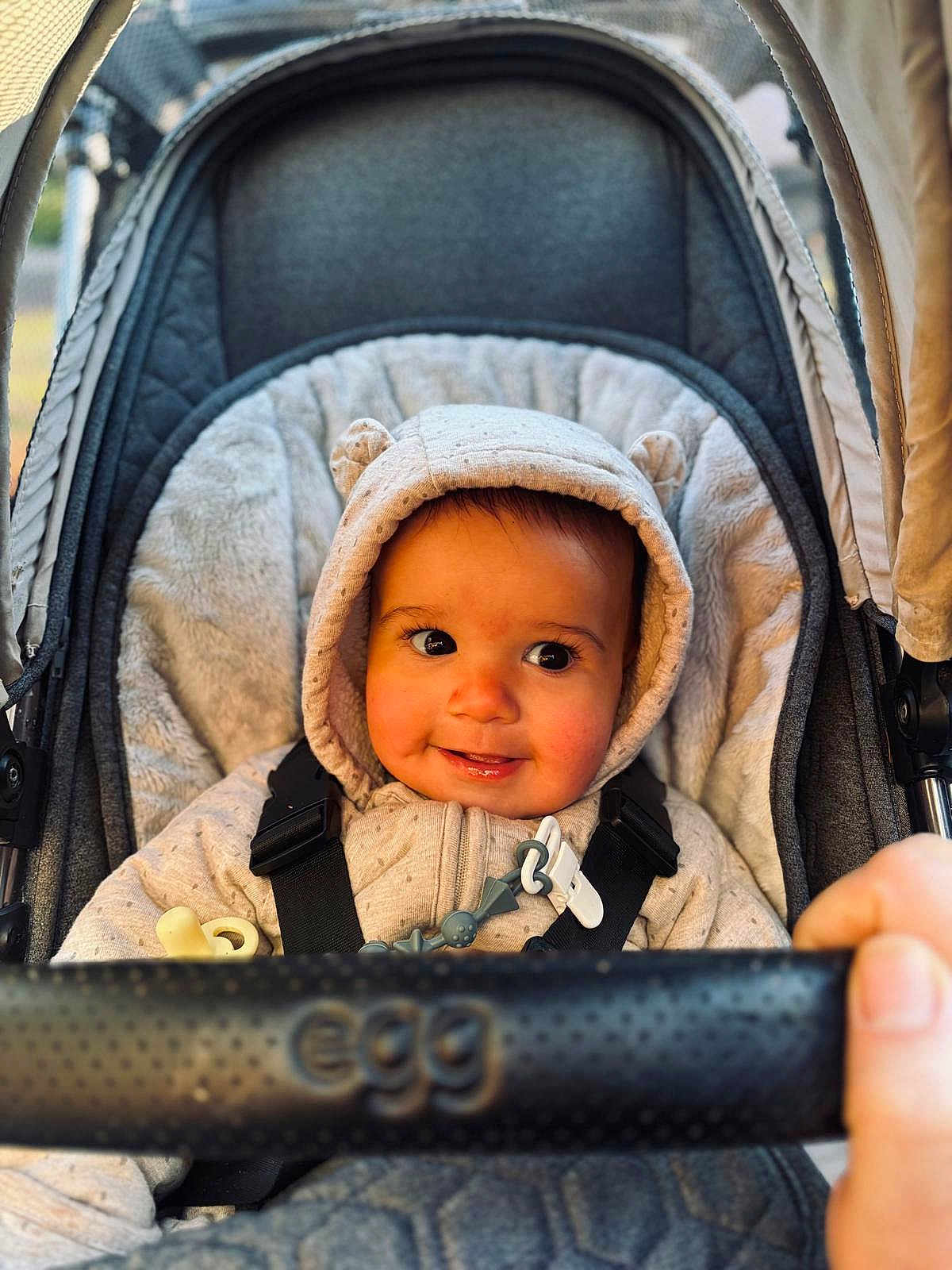 Otis is registered to the contest to win money with this photo: baby, child, stroller, hooded_jacket, smile, cute, face, infant, outdoor, hand, seat, straps, warm_clothing, plush, comfort, person, closeup, portrait, expression, baby_accessory