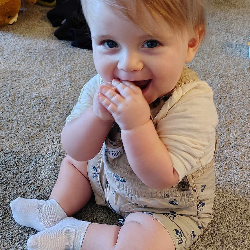 Hunter Allen is registered to the contest to win money with this photo: baby, child, toddler, sitting, smiling, happy, indoor, carpet, portrait, face, blue_eyes, blond_hair, white_socks, onesie, clothing, teddy_bear, toy_bag, home, floor, playful