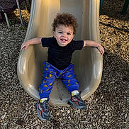 Remingtyn is registered to the contest to win money with this photo: toddler, child, smiling, curly_hair, slide, playground, outdoor, wood_chips, black_shirt, blue_pants, sneakers, happy, sitting, play, fun, daylight, park, recreation, person, casual_clothing