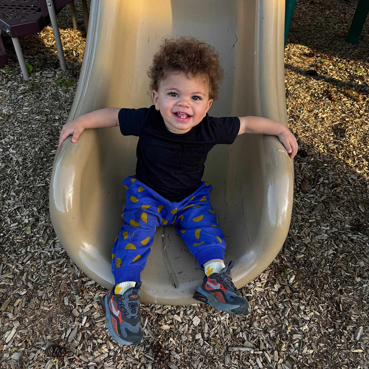 Remingtyn is registered to the contest to win money with this photo: toddler, child, smiling, curly_hair, slide, playground, outdoor, wood_chips, black_shirt, blue_pants, sneakers, happy, sitting, play, fun, daylight, park, recreation, person, casual_clothing