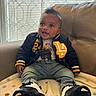 baby, blanket, casual, child, clothing, couch, cozy, face, footwear, happy, head, indoor, jacket, jeans, light, person, seated, smile, sneakers, window