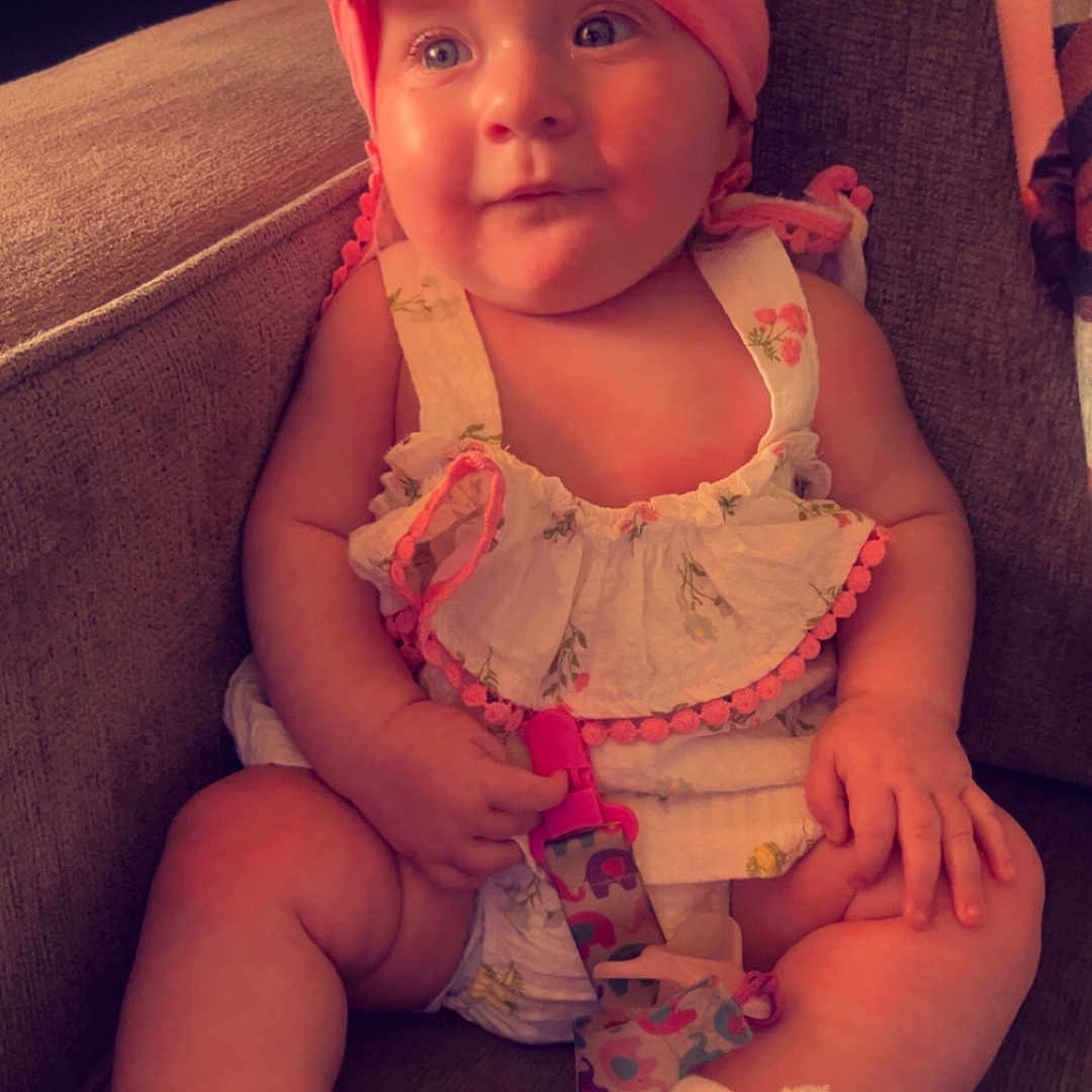 Willow is registered to the contest to win money with this photo: baby, child, chubby_arms, comfortable, couch, curious, cute, dress, expression, fabric, footwear, headband, indoors, infant, pacifier, pink, sitting, soft_lighting, toy, white_booties