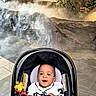 baby, car_seat, toy, smiling, indoor, rock_wall, waterfall, mist, plants, floor_tiles, happy, child, person, clothing, seated, cute, expression, leisure, playful, bright