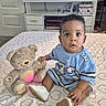 baby, teddy_bear, bed, bedspread, heart_pattern, blue_clothing, indoor, television, furniture, carpet, drawer, cabinet, toy, footwear, curly_hair, child, person, sitting, expression, room