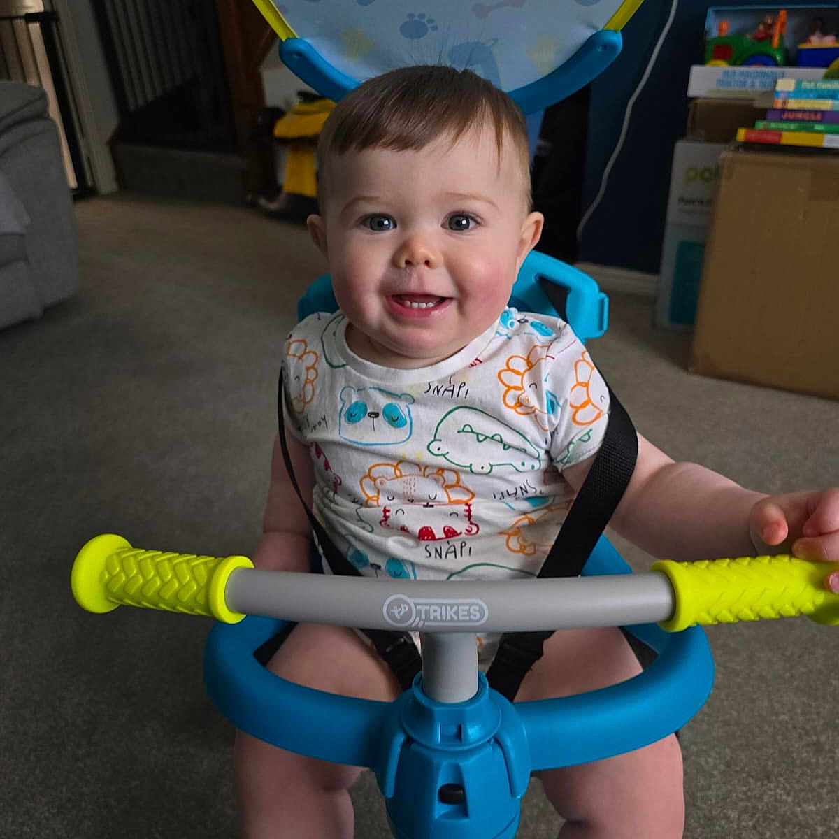 Luca is registered to the contest to win money with this photo: baby, blue, carpet, child, cute, face, furniture, handlebar, happy, indoor, living_room, pattern, person, playful, seat, shirt, smiling, toy, tricycle, yellow