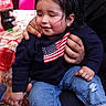american_flag, blue, bottle, child, clothing, coca_cola, cute, face, flag, hair, hand, holding, indoor, jeans, person, red, sitting, smile, sweater, toddler