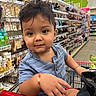 toddler, child, shopping_cart, supermarket, aisle, beauty_products, bracelet, blue_shirt, curly_hair, cute, face, person, retail, store, grocery, young_child, casual_clothing, indoors, playful, portrait