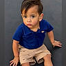 child, toddler, boy, sitting, blue_polo, beige_shorts, shorts, curious, expression, indoors, dark_background, cute, portrait, young_child, hands, face, hair, looking_up, casual_clothing, small
