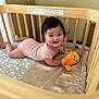 Olivia is registered to the contest to win money with this photo: baby, infant, crib, wooden_crib, plush_toy, orange_toy, smiling, happy, child, pink_outfit, lying_down, indoor, mattress, patterned_sheet, face, person, cute, young_child, toy, cozy