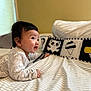 baby, infant, tummy_time, bed, striped_bedspread, pillow, cartoon_pillow, cute, child, indoors, smiling, black_hair, sleepwear, cozy, curious, young_child, soft_lighting, portrait, bedroom, resting
