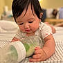 Olivia is registered to the contest to win money with this photo: baby, infant, bottle, milk, bed, striped_bedspread, lying_down, cute, chubby_cheeks, dark_hair, focus, indoor, soft_lighting, blurred_background, hand, clothing, short_sleeves, child, portrait, closeup
