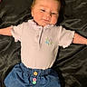 arms_outstretched, baby, black_background, buttons, child, clothing, cute, denim_skirt, face, floral_embroidery, happy, indoor, infant, lying_down, newborn, person, portrait, purple_top, smiling, soft_fabric