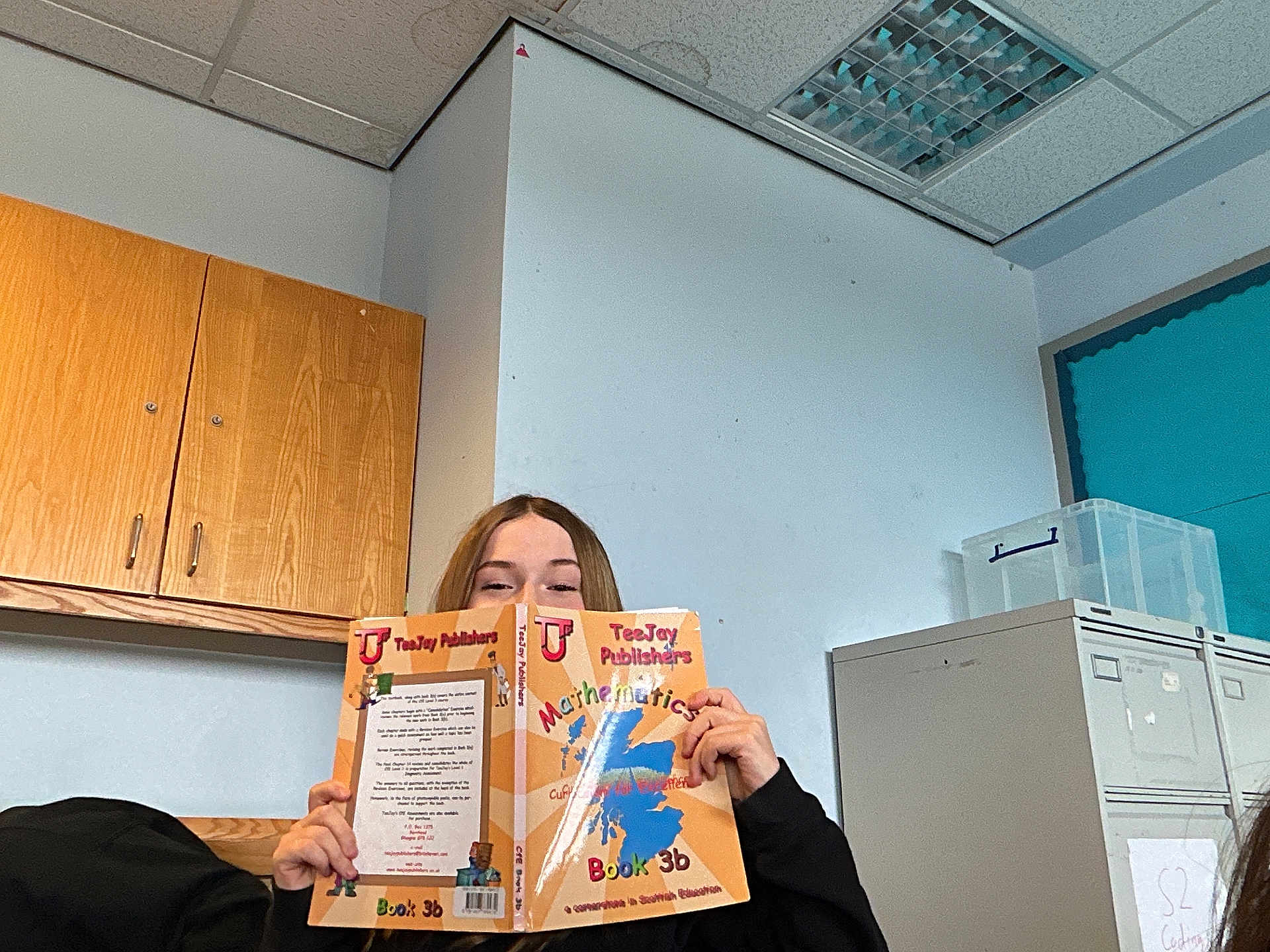 Butterfingers is registered to the contest to win money with this photo: child, book, reading, classroom, person, face, hands, wooden_cabinet, filing_cabinet, plastic_box, ceiling, fluorescent_light, bulletin_board, backpack, wall, desk, student, smile, sweater, book_cover