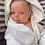 baby, child, towel, hooded_towel, tongue_out, playful, cute, striped_cushion, infant, face, skin, blue_eyes, blanket, wrapped, young, person, soft_texture, relaxed, indoors, smiling