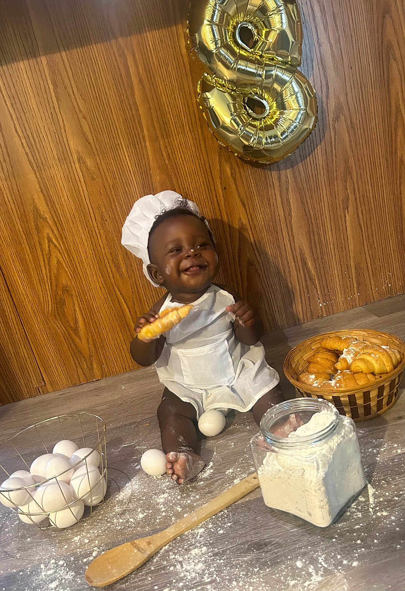 Cotie is registered to the contest to win money with this photo: baby, child, chef_hat, apron, croissant, eggs, flour, wooden_spoon, basket, floor, smiling, happy, baking, food, kitchen, balloon, celebration, portrait, indoor, cooking
