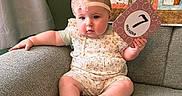 Riley joined the competition — help win amazing prizes! baby, infant, child, romper, headband, pink_bow, milestone_card, 7_months, couch, gray_furniture, quilt, patchwork, floral_pattern, sitting, indoors, curious_expression, chubby_cheeks, soft_light, cozy, cute