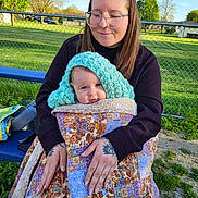 Riley joined the competition — help win amazing prizes! baby, woman, glasses, quilt, blue_hood, bench, outdoor, park, fence, grass, daylight, holding, smile, tattoo, casual_clothing, nature, person, portrait, child, family