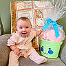 Riley is registered to the contest to win money with this photo: baby, child, infant, pink_outfit, smiling, armchair, quilt, patchwork, green_bucket, bunny_face, blue_bow, gift_wrap, home_interior, cute, happy, seated, indoor, soft_lighting, person, portrait