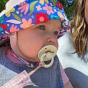 Riley joined the competition — help win amazing prizes! adult, baby, blue, child, closeup, clothing, face, floral_pattern, hat, headwear, nature, outdoor, pacifier, person, pink, portrait, summer, sunlight, trees, woman