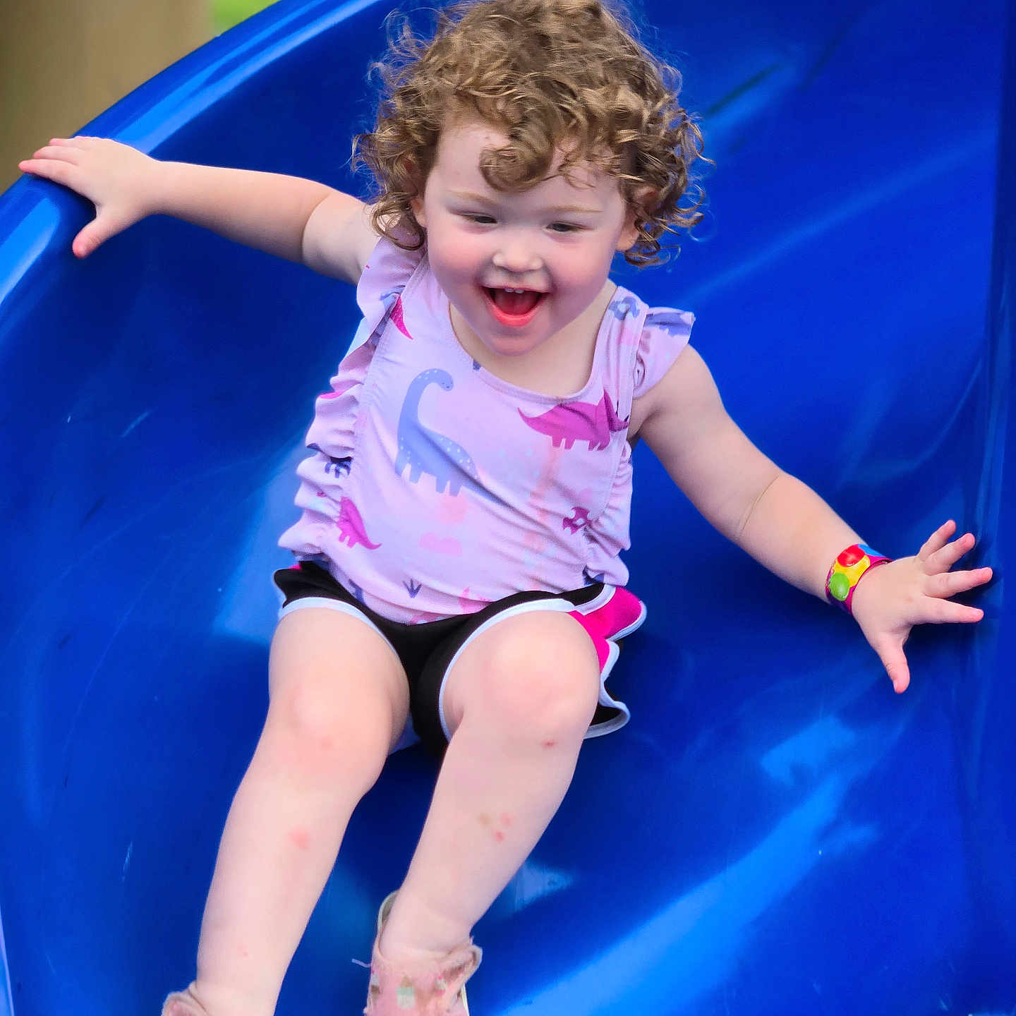 Cloie is registered to the contest to win money with this photo: active, arms_outstretched, blue_slide, casual_wear, child, curly_hair, cute, daylight, dinosaur_print, fun, happy, outdoor, park, pink_shoes, playground, playing, slide, smiling, summer_clothing, toddler