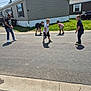 child, adult, street, road, grass, mobile_home, house, sidewalk, sunny, daytime, people, casual_clothing, game, outdoor, shoes, tshirt, shorts, jeans, greenery, dandelions