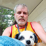Ellie is registered to the contest to win money with this photo: man, dog, outdoor, porch, tree, chair, casual, beard, curly_hair, yellow_tank_top, wood, pet, animal, face, blue_jeans, earrings, nature, relaxed, daylight, sitting