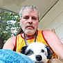 man, dog, outdoor, porch, tree, chair, casual, beard, curly_hair, yellow_tank_top, wood, pet, animal, face, blue_jeans, earrings, nature, relaxed, daylight, sitting