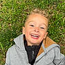 child, boy, smiling, happy, laughing, portrait, outdoor, grass, lying_down, face, teeth, blond_hair, jacket, hoodie, zipper, nature, garden, close_up, daylight, casual_clothing