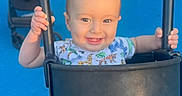 Patrick joined the competition — help win amazing prizes! baby, child, toddler, smiling, happy, swing, playground, outdoor, seated, infant, face, portrait, hands, chains, bucket_seat, blue_floor, white_fence, stroller, greenery, photography