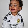 child, toddler, soccer_ball, real_madrid, jersey, smiling, happy, portrait, indoor, short_hair, white_background, sportswear, football, cute, young, person, hands, face, eyes, clothing