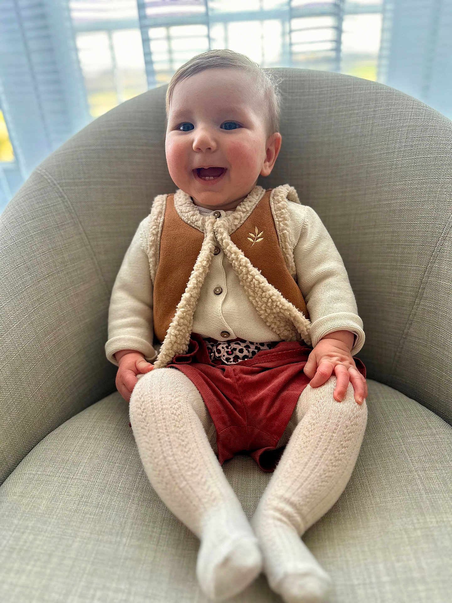 Céleste a rejoint le concours — aidez-le/la à gagner de superbes lots ! baby, child, smile, happy, armchair, clothing, vest, knitted_tights, tights, indoor, person, infant, cute, face, sitting, window, natural_light, cozy, warm_clothing, blinds