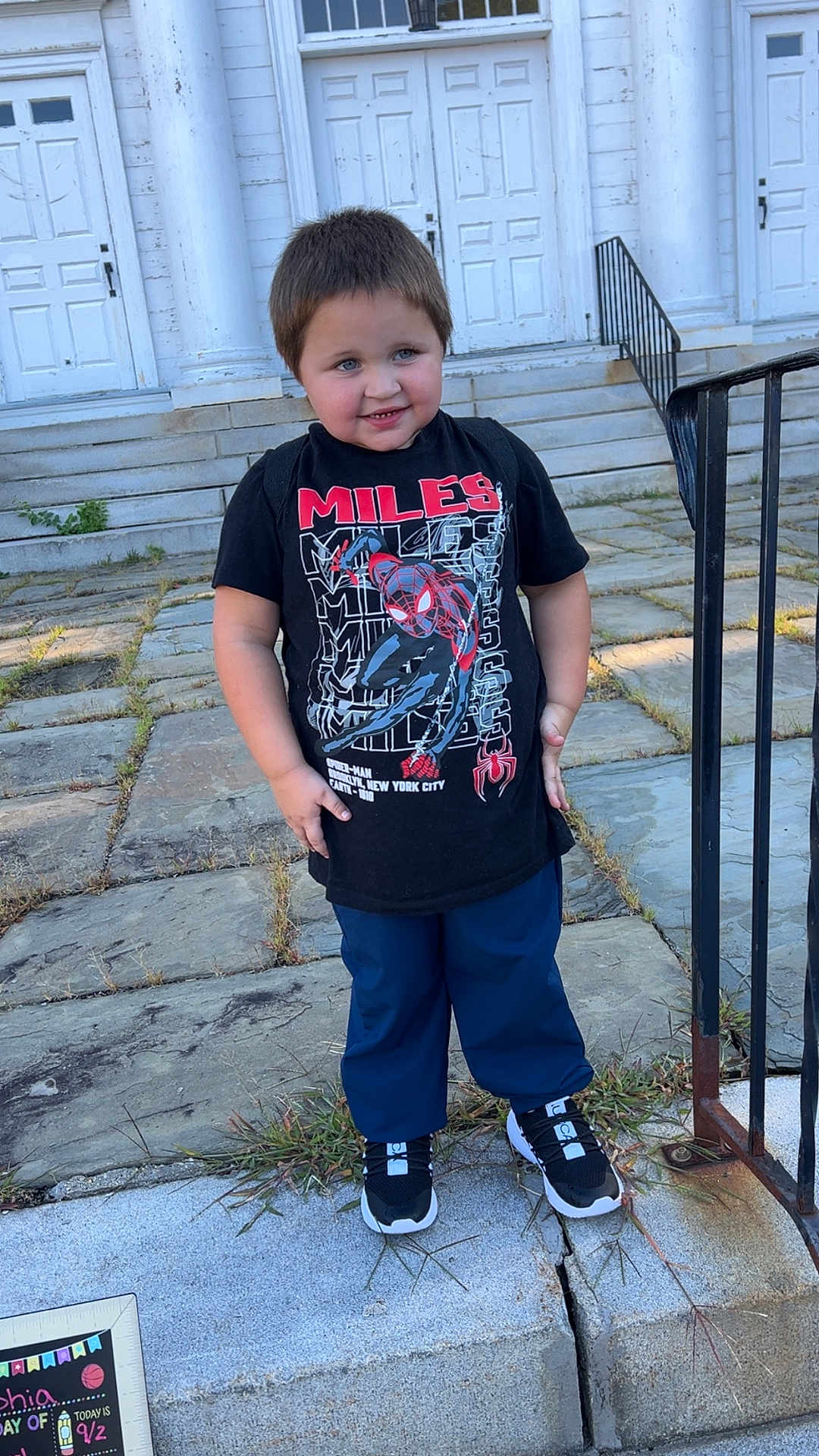 Malachi joined the competition — help win amazing prizes! child, boy, standing, smiling, black_tshirt, spiderman, blue_pants, sneakers, sidewalk, steps, white_building, door, railing, outdoor, grass, concrete, casual_clothing, daylight, portrait, person