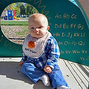 Aiden is registered to the contest to win money with this photo: baby, child, infant, playground, outdoor, sitting, portrait, face, smiling, blue_clothing, checkered_shirt, bib, white_socks, sunlight, alphabet, play_structure, slide, grass, fence, daytime