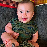 Maverick is registered to the contest to win money with this photo: baby, infant, child, smiling, happy, sitting, couch, blanket, leopard_print, camouflage, clothing, foot, barefoot, portrait, indoor, home, pillow, face, eyes, joy