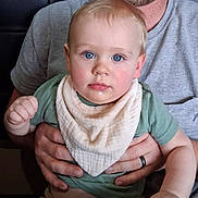 Miles is registered to the contest to win money with this photo: baby, adult, hands, blue_eyes, bib, green_shirt, smiling_adult, facial_hair, indoor, table, curious_expression, close_up, portrait, child, person, skin, human, seated, holding, caucasian