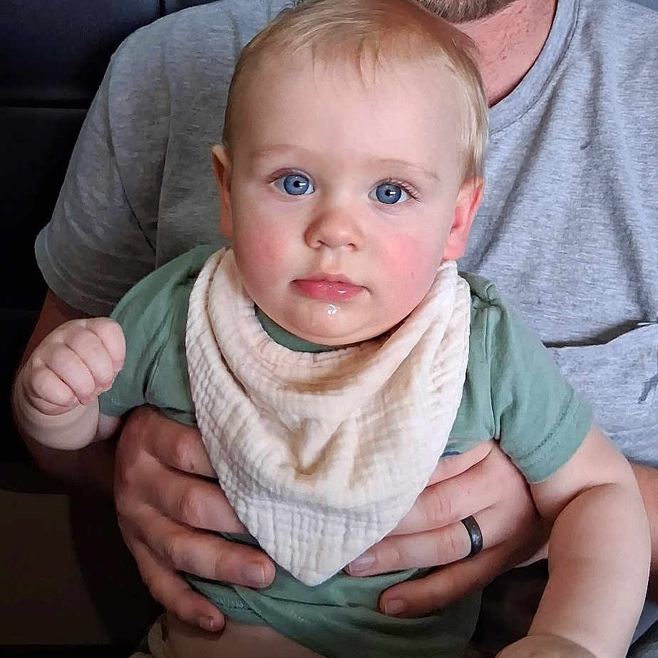 Miles is registered to the contest to win money with this photo: baby, adult, hands, blue_eyes, bib, green_shirt, smiling_adult, facial_hair, indoor, table, curious_expression, close_up, portrait, child, person, skin, human, seated, holding, caucasian