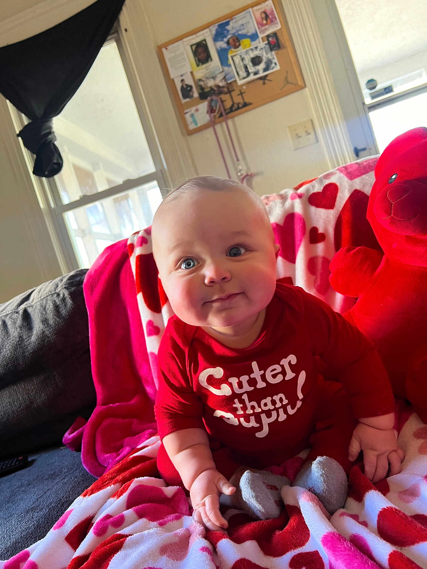 Zaylen is registered to the contest to win money with this photo: baby, child, red_clothing, teddy_bear, blanket, heart_pattern, couch, indoor, smiling, cute, portrait, person, infant, soft_toys, home, cozy, face, sitting, window, furniture