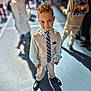 child, boy, suit, tie, vest, dress_shoes, curly_hair, indoor, event, crowd, portrait, formal_wear, smile, young_child, shallow_depth_of_field, spotlight, floor, shadow, people, blurred_background