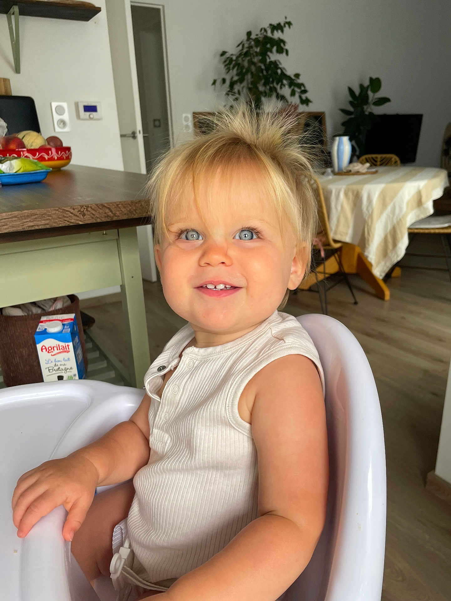 Eïra a rejoint le concours — aidez-le/la à gagner de superbes lots ! toddler, child, high_chair, blonde_hair, blue_eyes, smiling, indoor, kitchen, table, chair, fruit_bowl, milk_carton, wooden_floor, plant, white_clothing, happy, portrait, baby, furniture, home