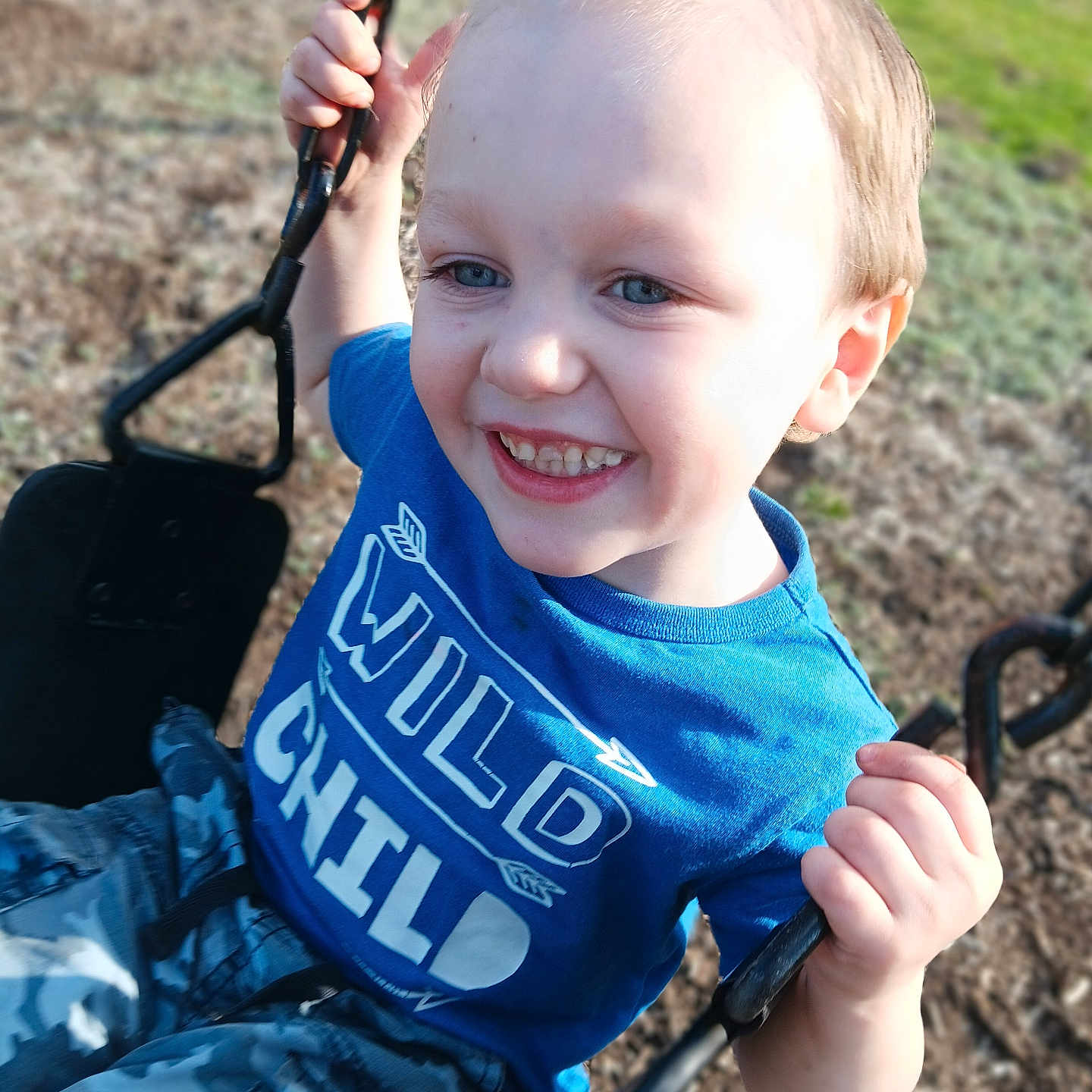 Teagen is registered to the contest to win money with this photo: blue_tshirt, boy, camouflage_pants, candid, chain, child, close_up, grass, holding_chain, outdoors, playground, playtime, portrait, seat, shirt_text, smiling, spiky_hair, sunlight, swing, teeth