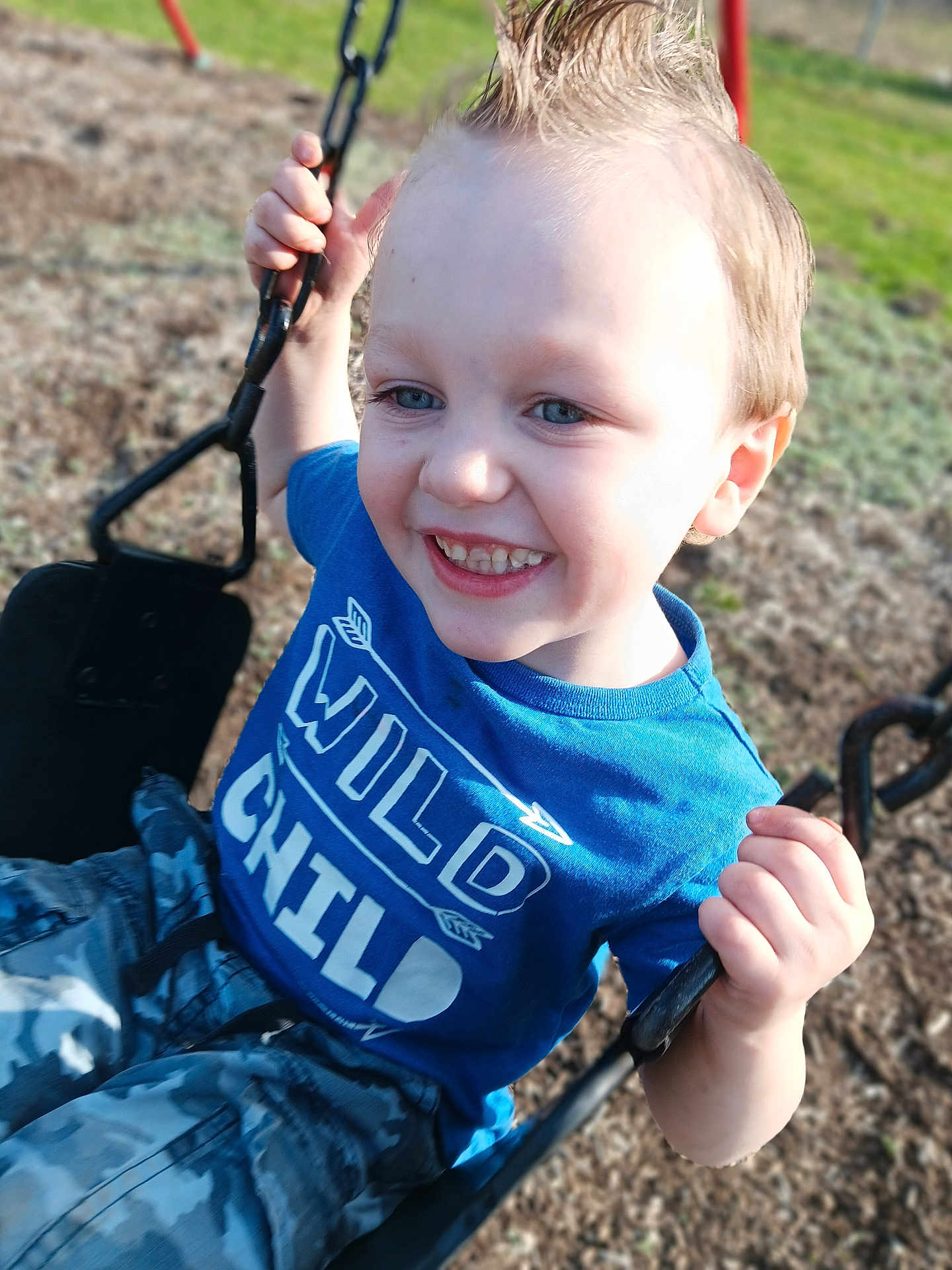 Teagen is registered to the contest to win money with this photo: child, boy, smiling, swing, playground, outdoors, grass, blue_tshirt, shirt_text, camouflage_pants, close_up, portrait, spiky_hair, teeth, holding_chain, seat, chain, candid, sunlight, playtime