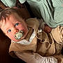 baby, pacifier, blue_eyes, beige_vest, white_shirt, tattoo, soft_fabric, green_fabric, infant, close_up, portrait, child, cute, indoors, relaxed, hand, face, clothing, person, skin